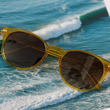 Peepers Boho II Sunglasses in translucent honey yellow resting on a rock with ocean waves behind