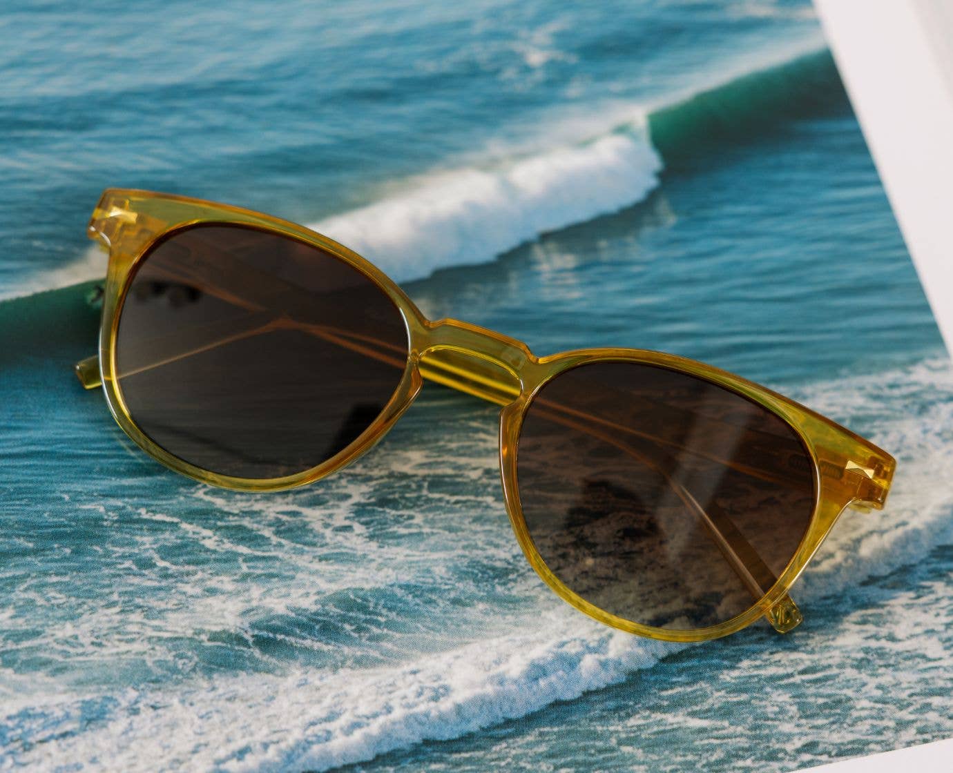 Peepers Boho II Sunglasses in translucent honey yellow resting on a rock with ocean waves behind