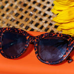 Peepers Capri Sunglasses in Leopard Tortoise resting on an orange surface with a woven hat and yellow flowers