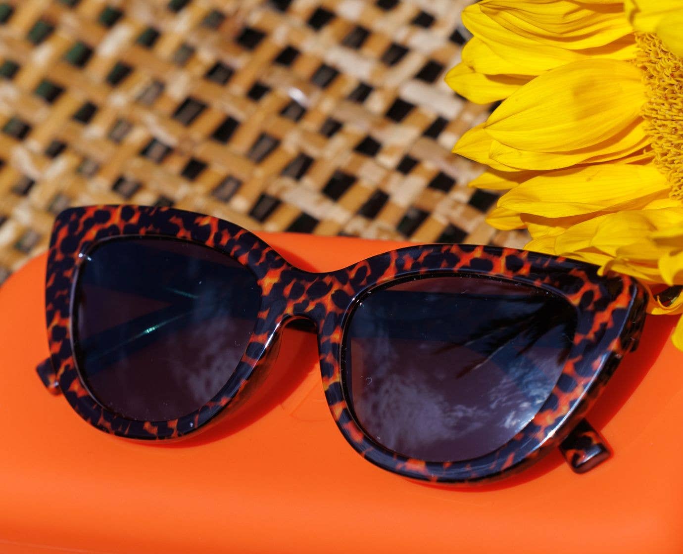 Peepers Capri Sunglasses in Leopard Tortoise resting on an orange surface with a woven hat and yellow flowers