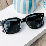 Peepers Clubhouse Sunglasses in Black resting on a book beside a blue striped vase