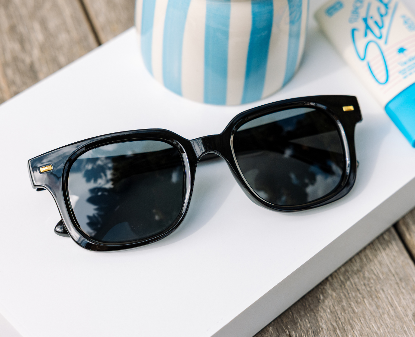 Peepers Clubhouse Sunglasses in Black resting on a book beside a blue striped vase