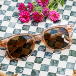 Peepers Clubhouse Sunglasses in Blush on a green checkered surface with pink flowers
