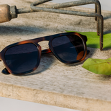 Peepers Deep Dive Sunglasses in Mocha Tortoise resting on a stone surface beside garden tools