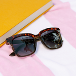 Peepers Frenchy Sunglasses in Green/Leopard Tortoise on a pink striped towel beside a yellow book