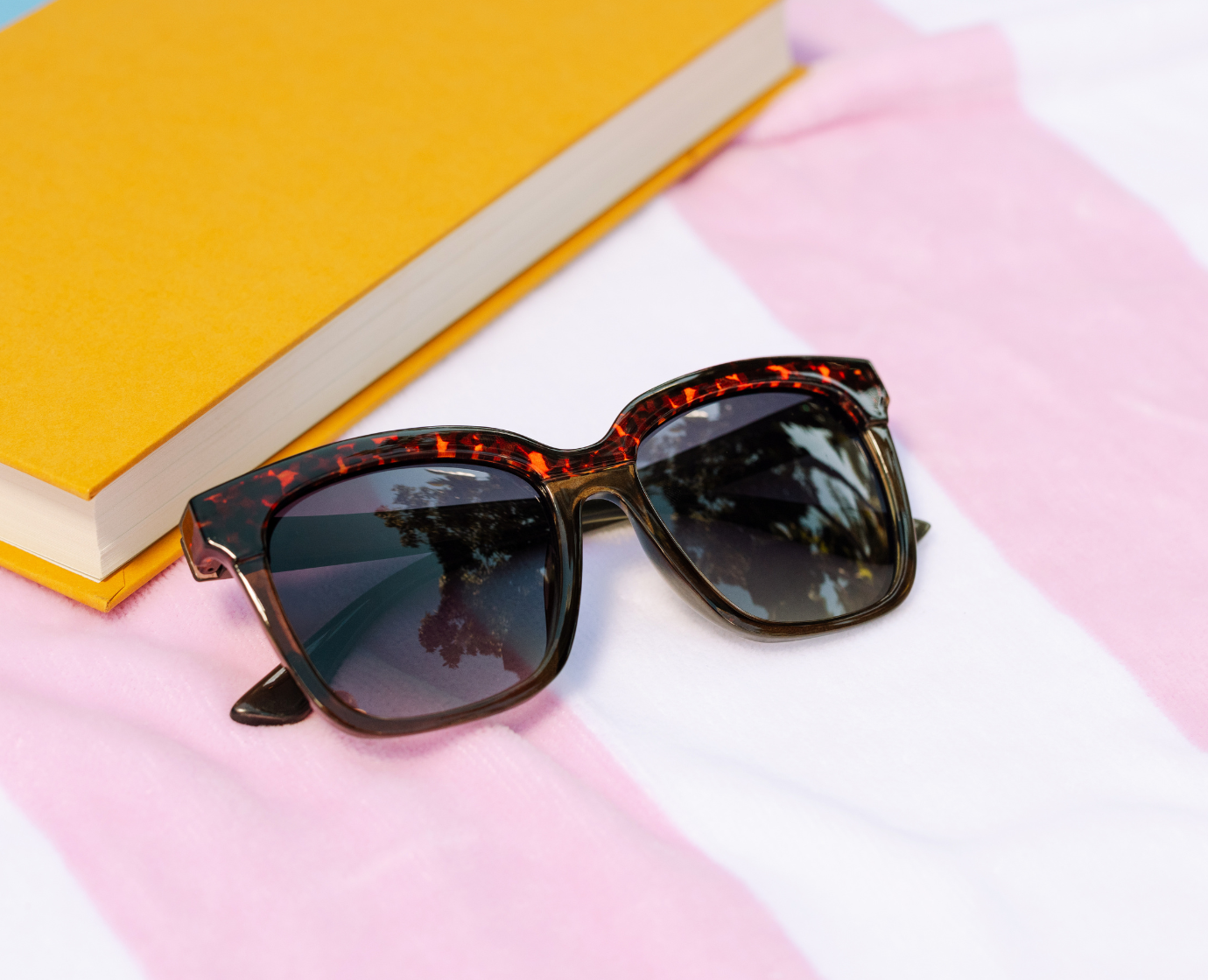 Peepers Frenchy Sunglasses in Green/Leopard Tortoise on a pink striped towel beside a yellow book