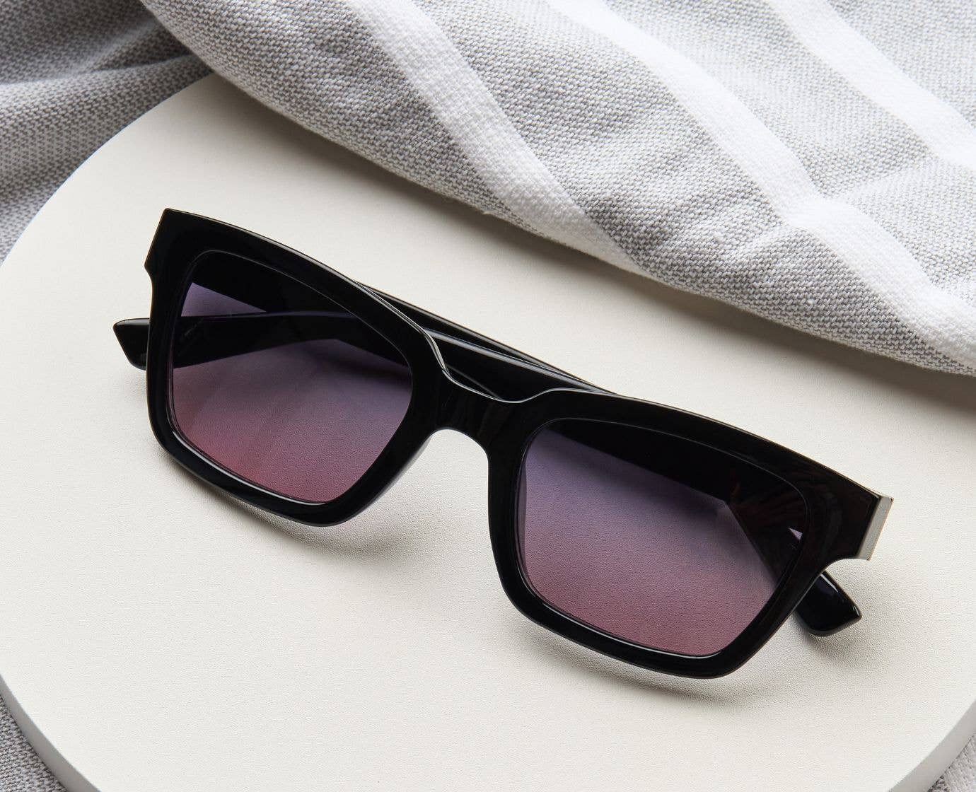 Peepers Myrtle Sunglasses in Black resting on a white surface beside a towel