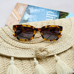 Peepers Myrtle Sunglasses in tortoise resting on a woven straw bag with a magazine