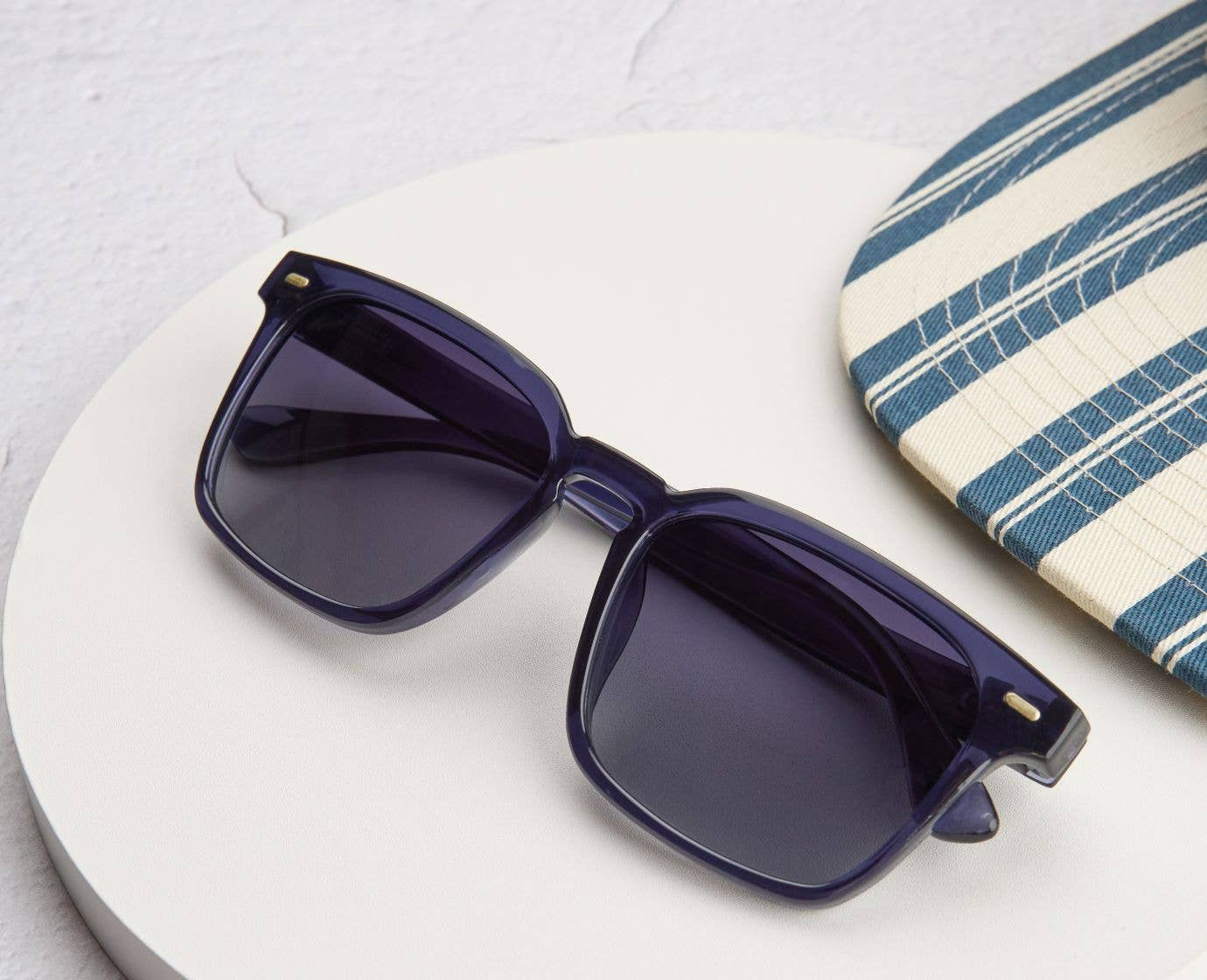 Peepers Shasta Sunglasses in Blue resting on a white table beside a striped surfboard