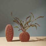 A short round Poppy Budvase holding green plant and a tall oval one in terracotta geometric silhouette with imprint of live plant. Muted dark background.