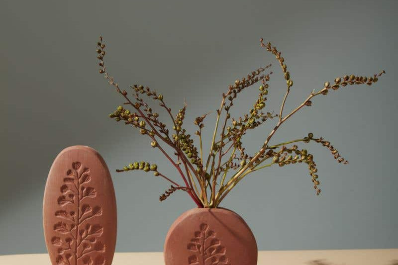 A short round Poppy Budvase holding green plant and a tall oval one in terracotta geometric silhouette with imprint of live plant. Muted dark background.