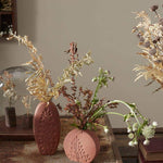 Tall oval and short round Poppy Budvases holding dried leafy plants. Set on basket table in home surrounded by other dried plants. 