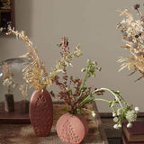 Tall oval and short round Poppy Budvases holding dried leafy plants. Set on basket table in home surrounded by other dried plants. 