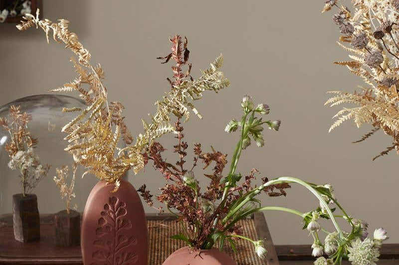 Tall oval and short round Poppy Budvases holding dried leafy plants. Set on basket table in home surrounded by other dried plants. 