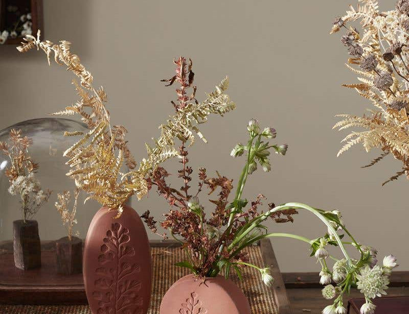 Tall oval and short round Poppy Budvases holding dried leafy plants. Set on basket table in home surrounded by other dried plants. 