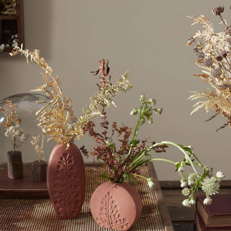 Tall oval and short round Poppy Budvases holding dried leafy plants. Set on basket table in home surrounded by other dried plants. 
