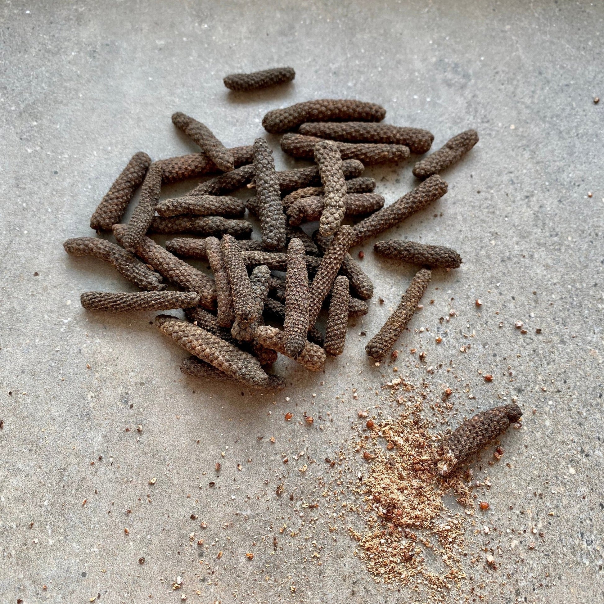 Rivsalt Java Long Pepper spikes scattered on a stone surface with ground pepper