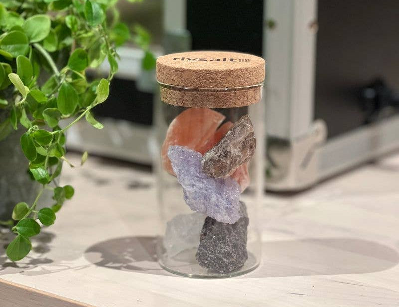 Rivsalt Salt Rock Selection Glass Jar with cork lid holding pink, purple and gray salt rocks on a wood table by a plant