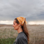 Woman in a field wearing the rust Rooted & Wild Adventure Co. Burn and Bloom Bandana as a head scarf, side view