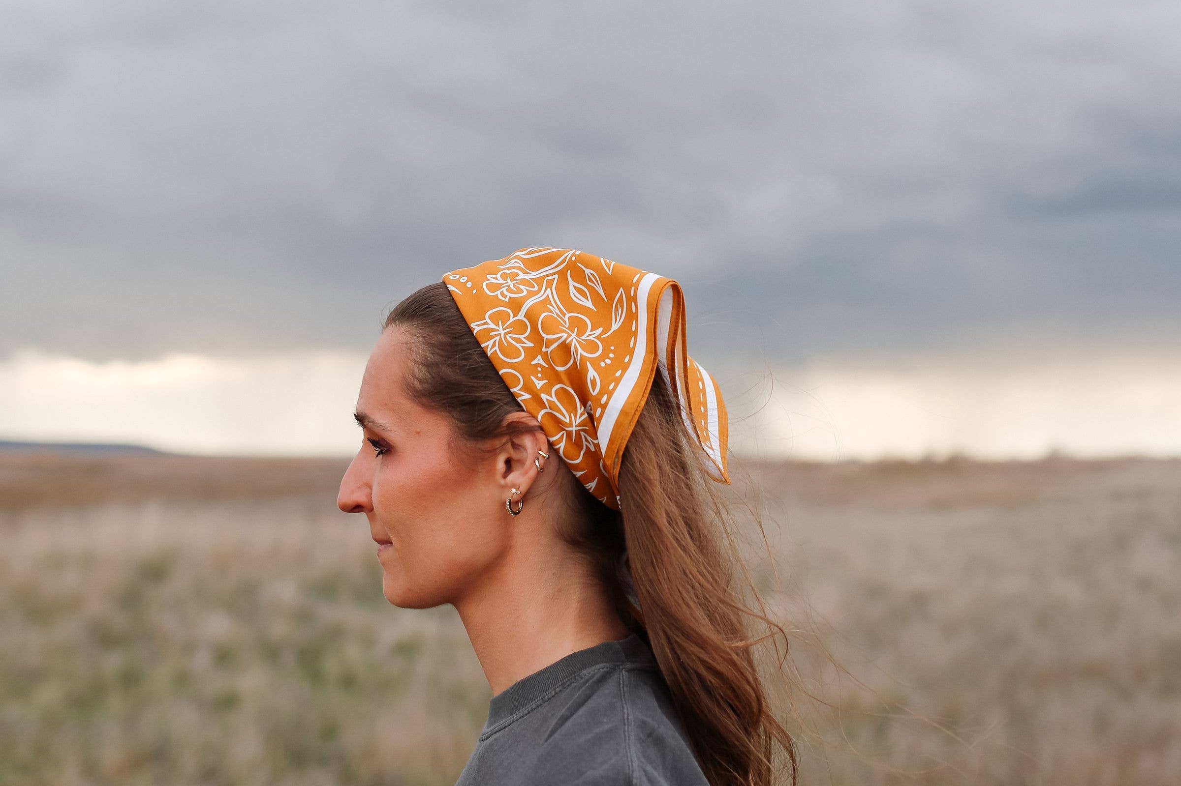 Woman in a field wearing the rust Rooted & Wild Adventure Co. Burn and Bloom Bandana as a head scarf, side view