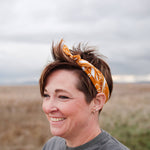 Smiling woman with short hair wearing the Rooted & Wild Adventure Co. Burn and Bloom Bandana as a headband