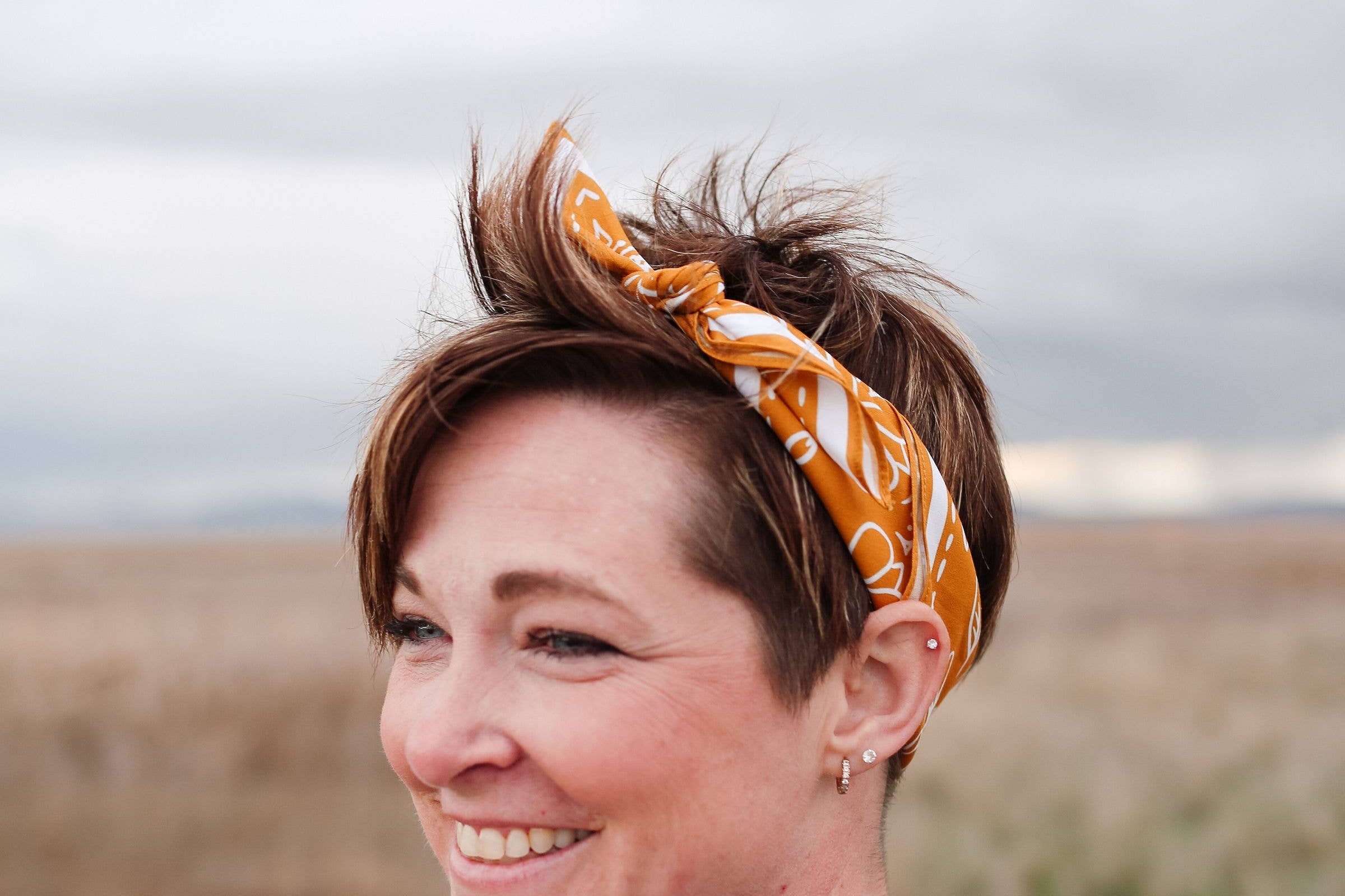 Smiling woman with short hair wearing the Rooted & Wild Adventure Co. Burn and Bloom Bandana as a headband