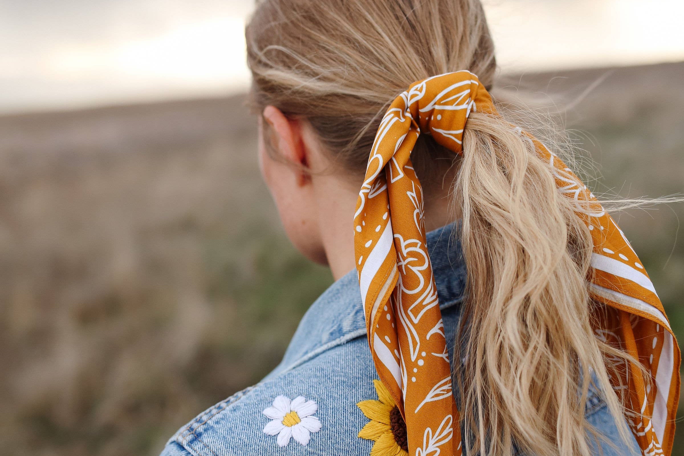 Rooted & Wild Adventure Co. Burn and Bloom Bandana tied around a blonde ponytail, worn with a denim jacket