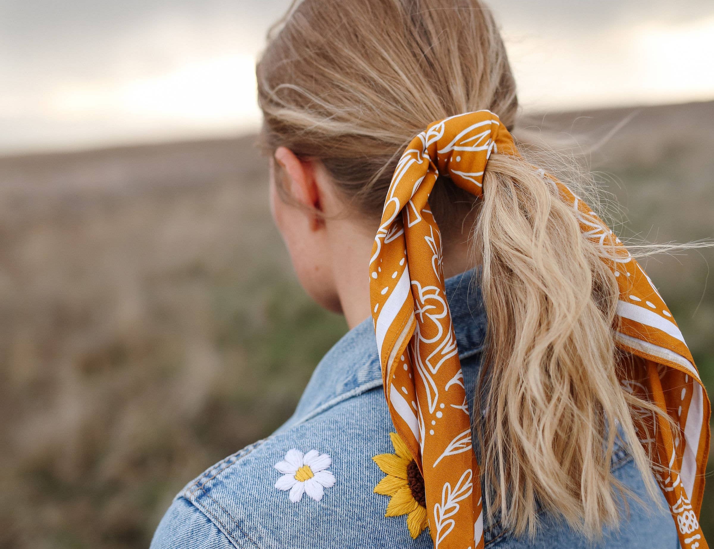 Rooted & Wild Adventure Co. Burn and Bloom Bandana tied around a blonde ponytail, worn with a denim jacket