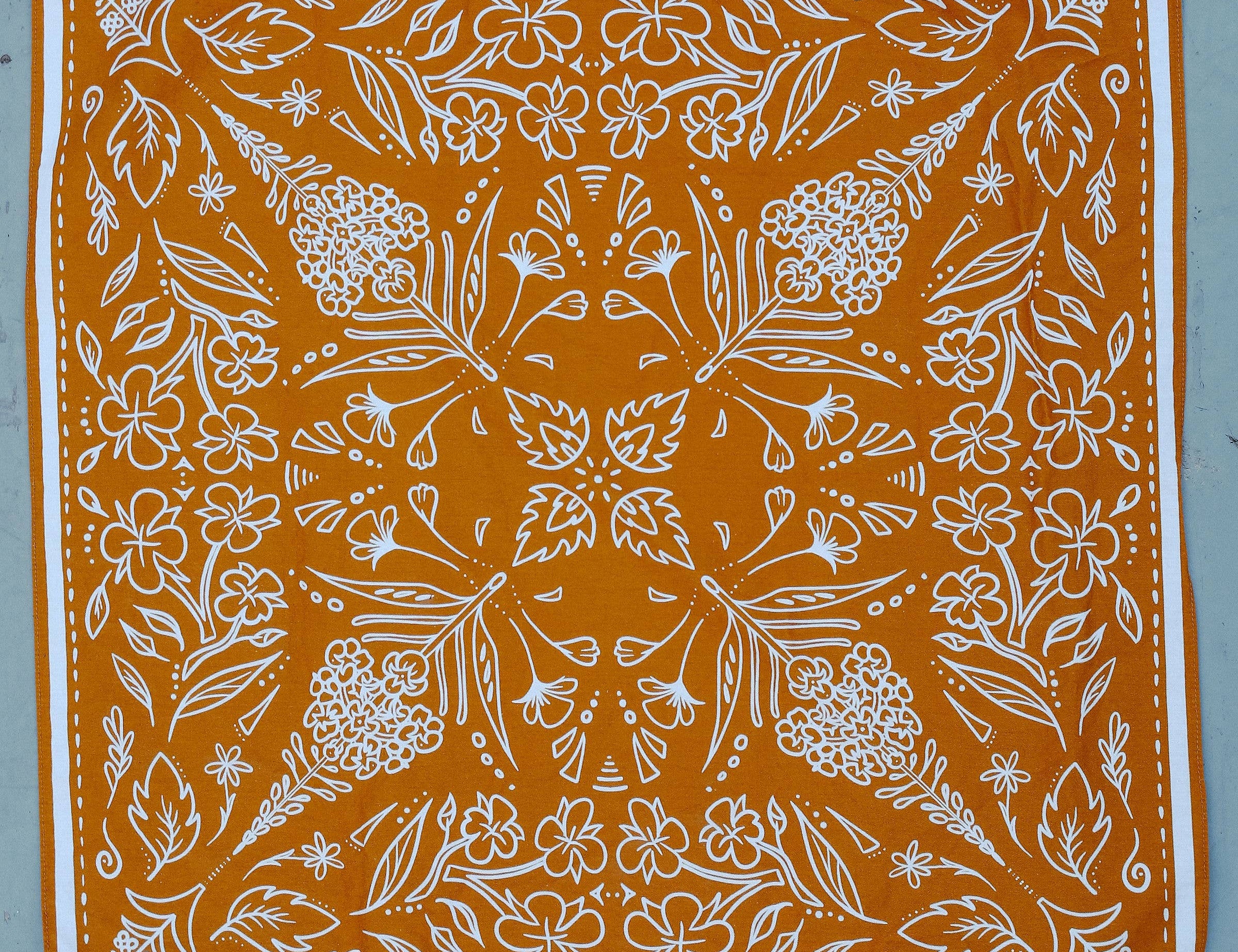 Rooted & Wild Adventure Co. Burn and Bloom Bandana, a rust orange bandana with a white wildflower print, laid flat