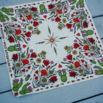 Rooted & Wild Adventure Co. Desert Bloom Bandana, a cream bandana with red cactus flowers and green botanicals, laid flat on blue wood