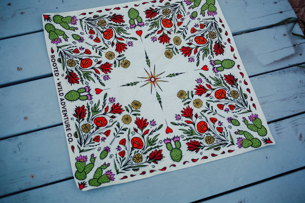 Rooted & Wild Adventure Co. Desert Bloom Bandana, a cream bandana with red cactus flowers and green botanicals, laid flat on blue wood