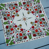 Rooted & Wild Adventure Co. Desert Bloom Bandana, a cream bandana with red cactus flowers and green botanicals, laid flat on blue wood
