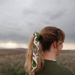 Rooted & Wild Adventure Co. Desert Bloom Bandana tied around a blonde ponytail under a stormy sky