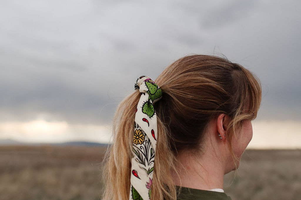 Rooted & Wild Adventure Co. Desert Bloom Bandana tied around a blonde ponytail under a stormy sky