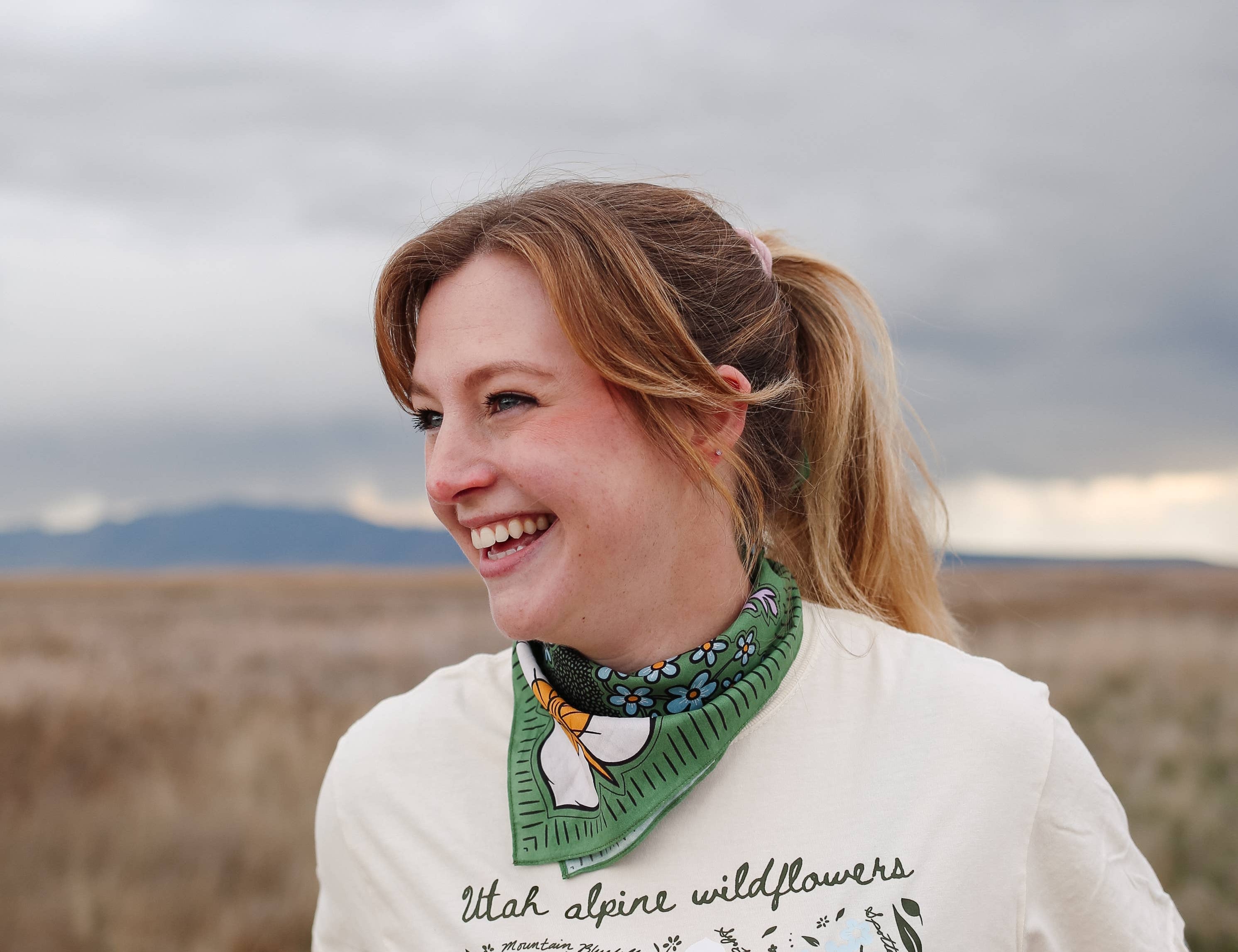 Smiling woman in a wildflower tee wearing the Rooted & Wild Adventure Co. High Meadows Bandana around her neck
