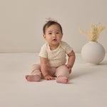 Rylee & Cru Baby Pant in Blush worn by a seated baby in a cream top beside a vase of dried grass