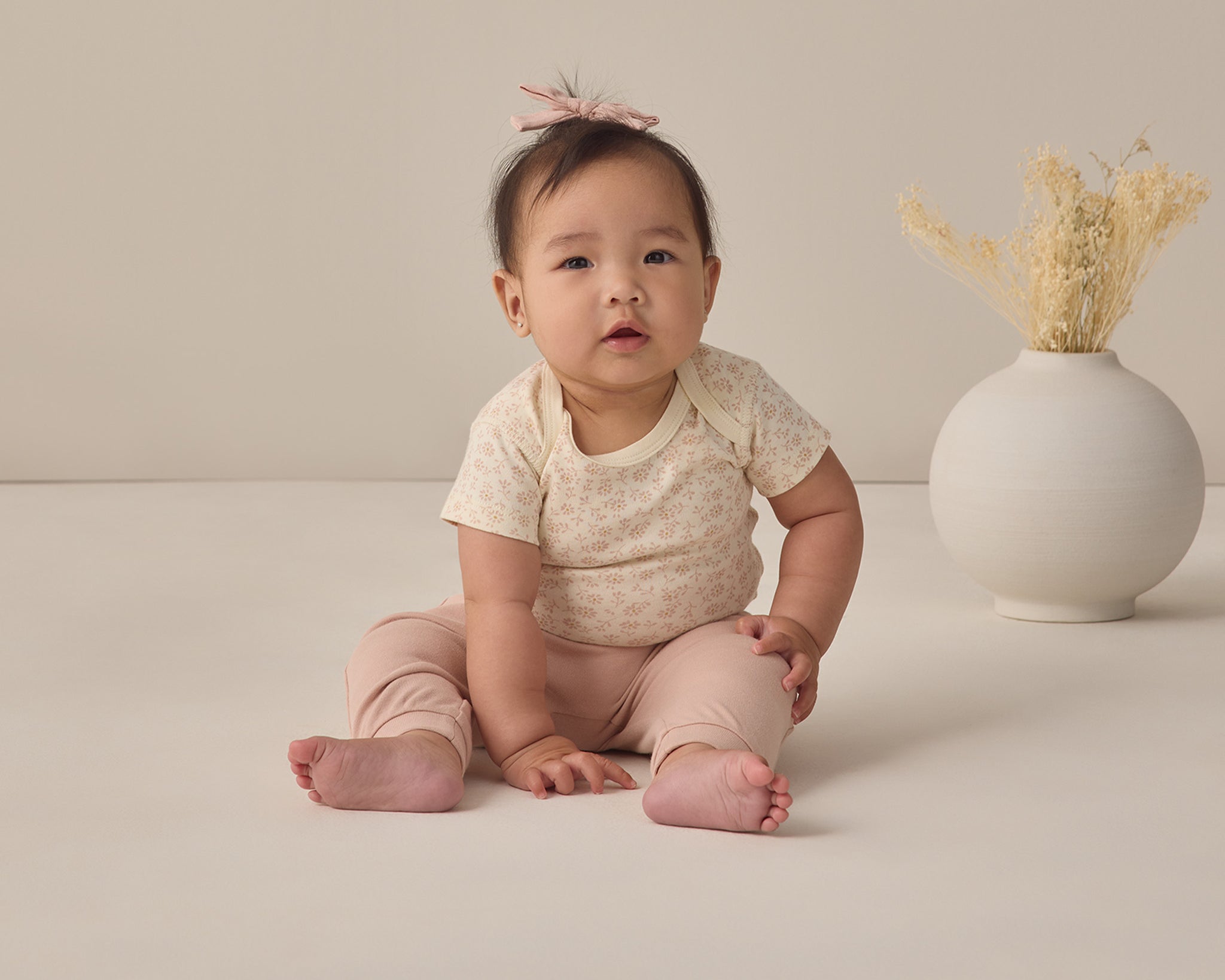 Rylee & Cru Baby Pant in Blush worn by a seated baby in a cream top beside a vase of dried grass