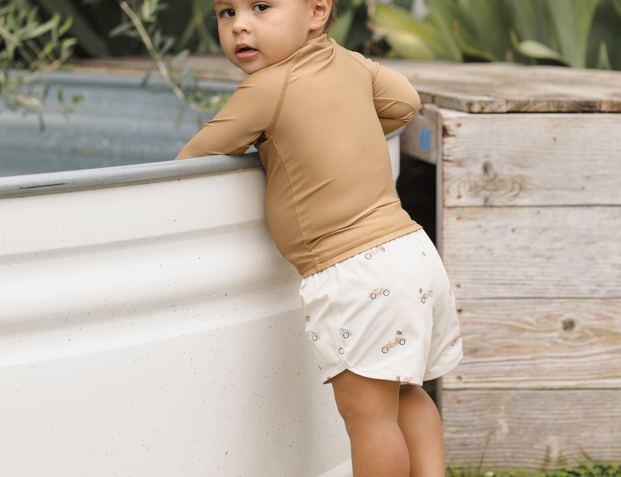 Toddler in a tan rash guard and Rylee & Cru Boys Swim Short in Race Car leaning on a stock tank pool