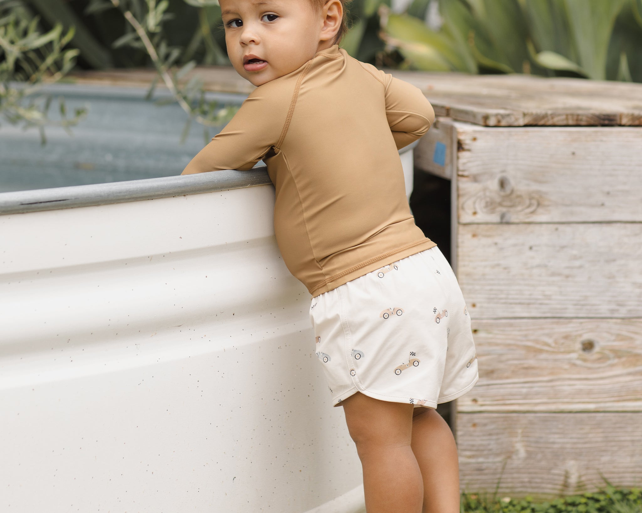 Toddler in a tan rash guard and Rylee & Cru Boys Swim Short in Race Car leaning on a stock tank pool