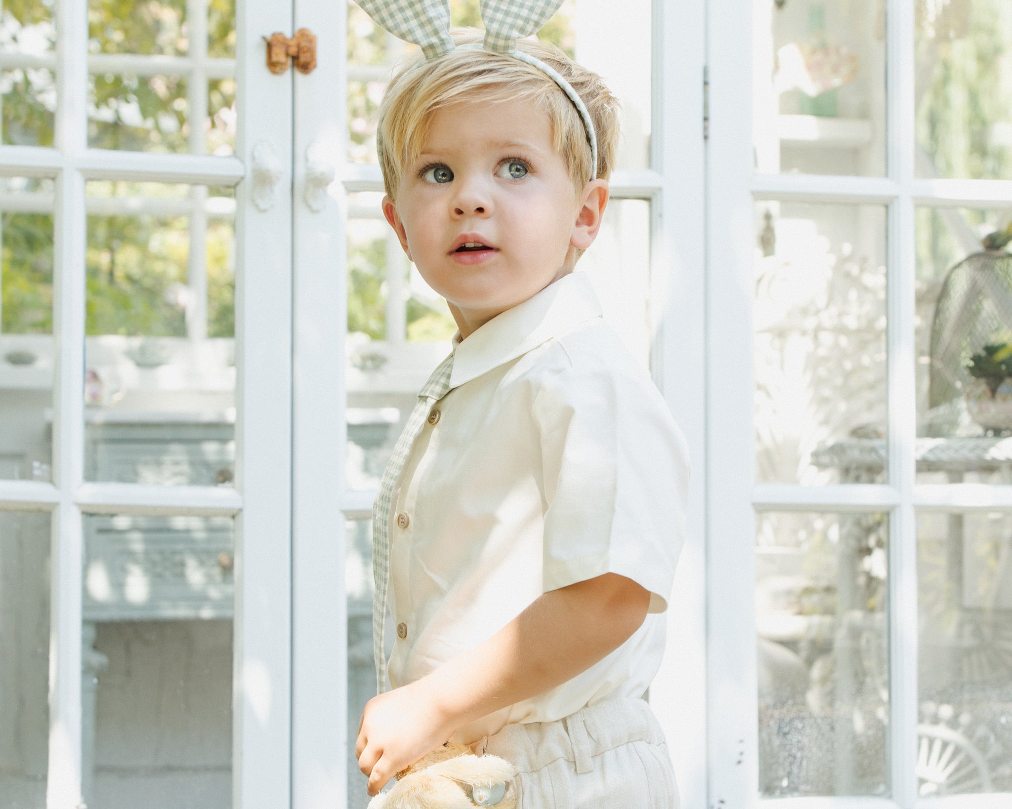 Boy in a white outfit wearing the Rylee & Cru Bunny Headband in Eucalyptus Check outside a greenhouse