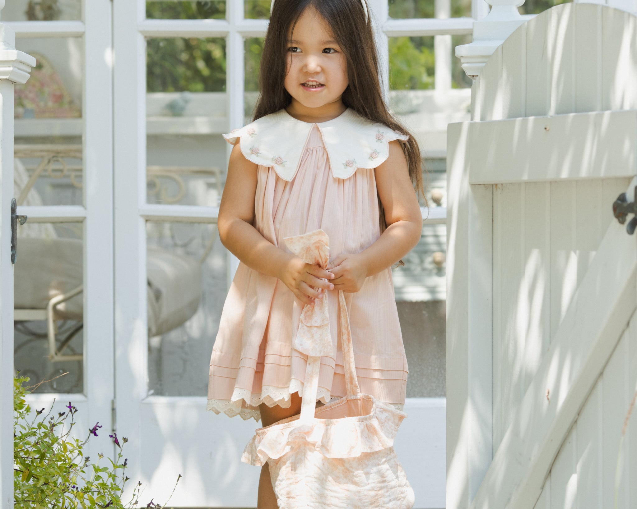 Girl in a pink dress wearing the Rylee & Cru Bunny Headband in Petal by a garden gate