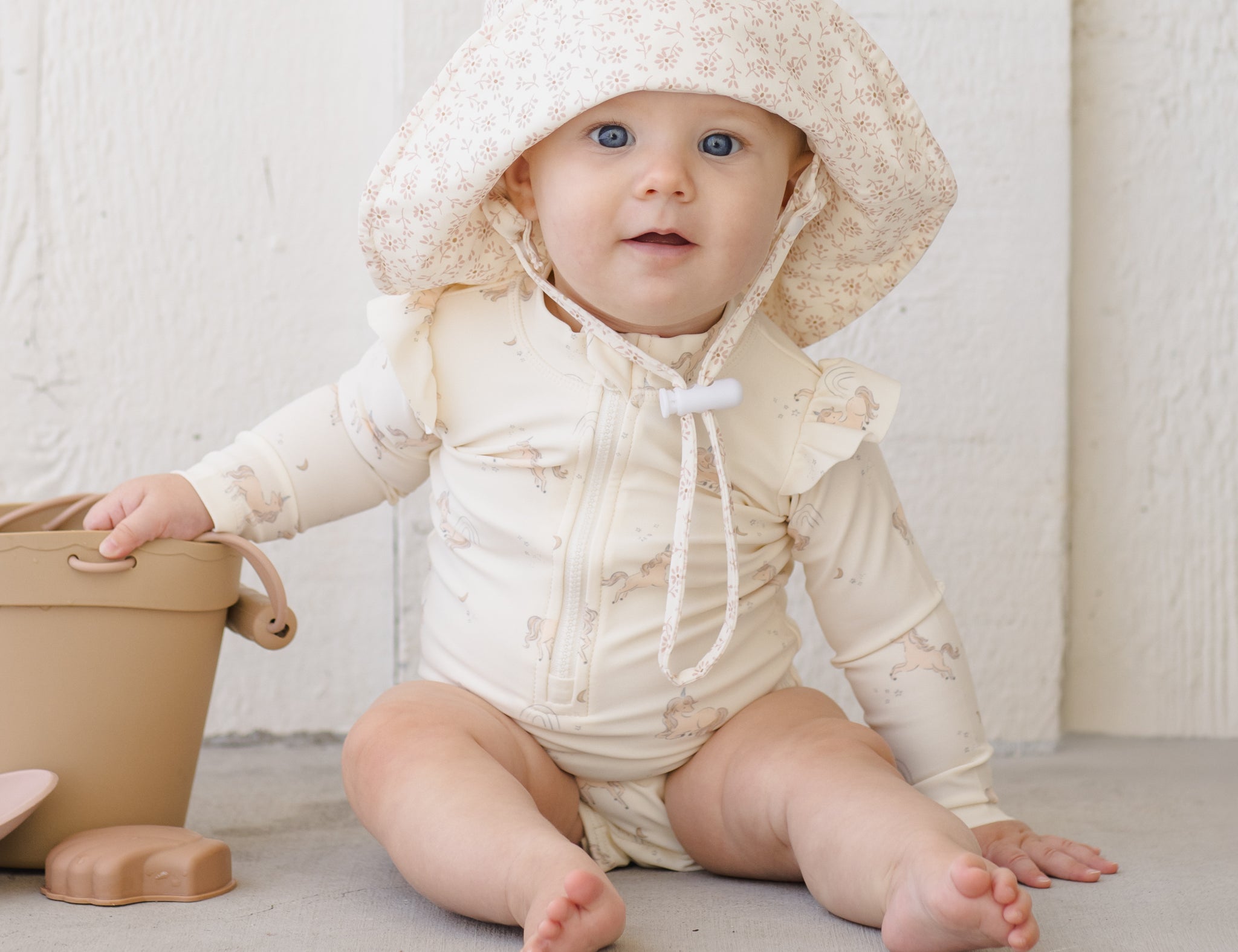 Baby in a sun hat wearing the Rylee & Cru Byron Rashguard One-Piece in Unicorns, playing with a toy bucket