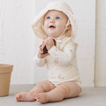 Baby in a sun hat and the cream Rylee & Cru Byron Rashguard One-Piece in Unicorns, sitting and smiling