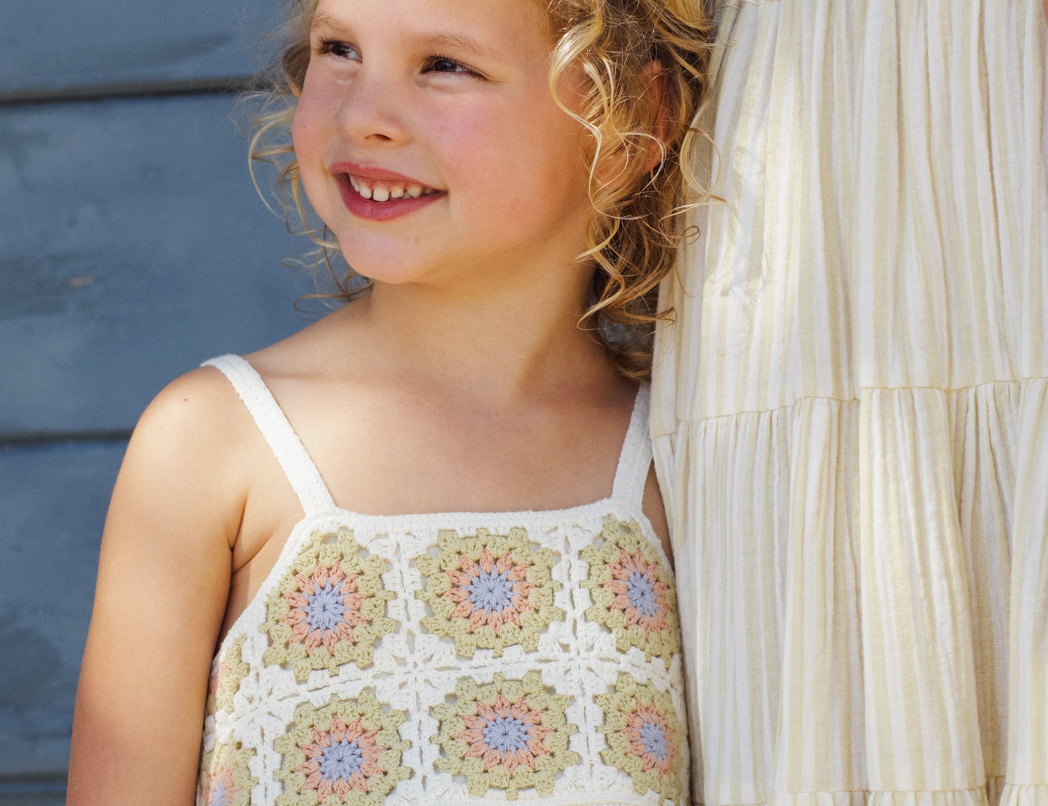Smiling girl leaning on a blue wall wearing the Rylee & Cru Crochet Sundress in Citron Daisy Check