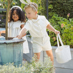 Boy in a white outfit carrying the Rylee & Cru Easter Basket in Sky Stripe beside a girl at a garden planter