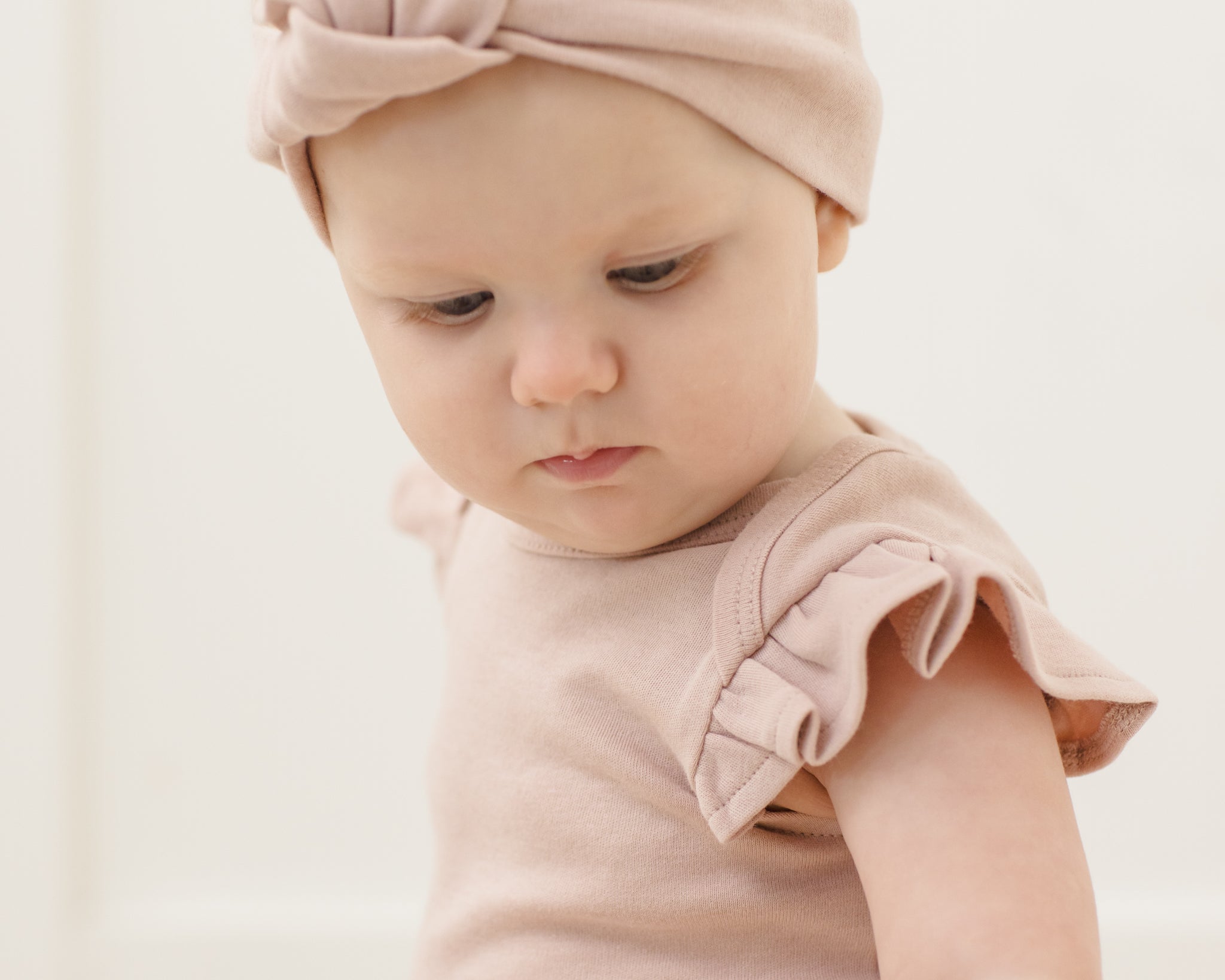 Close-up of a baby in a pink headband wearing the Rylee & Cru Flutter Sleeve Bodysuit in Blush
