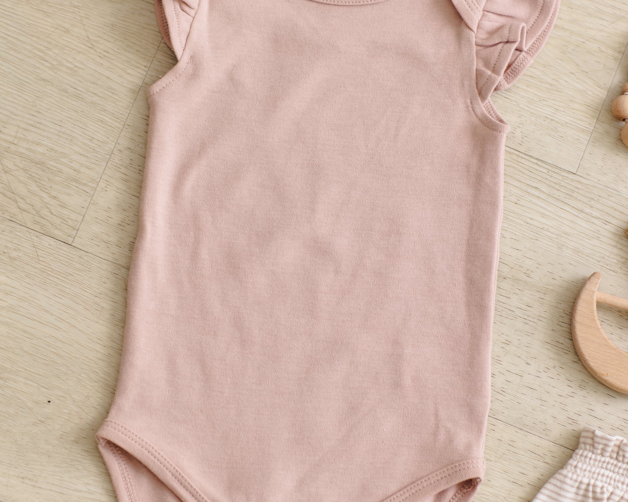 Rylee & Cru Flutter Sleeve Bodysuit in Blush laid on a wood floor with a wooden toy
