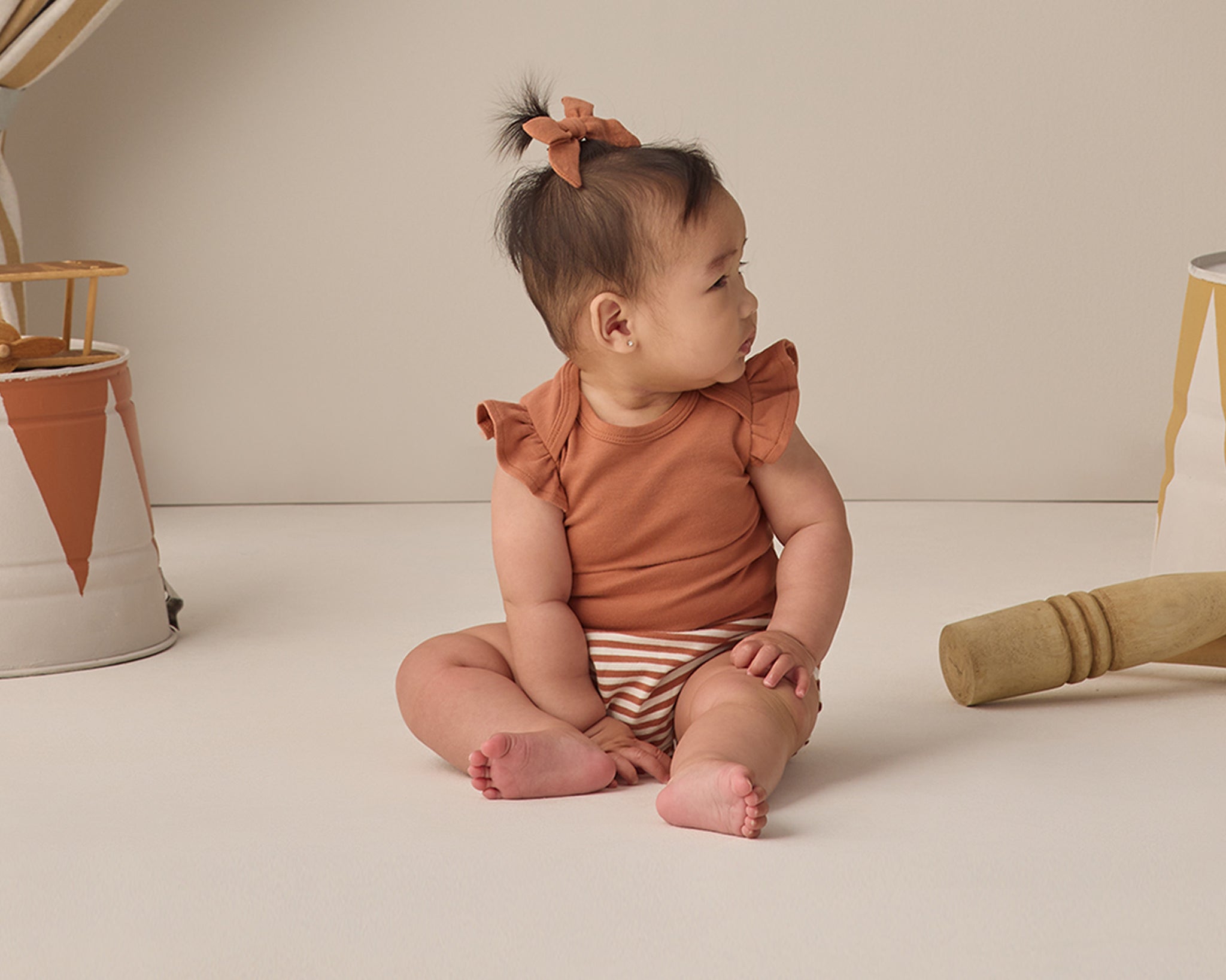 Baby with a hair bow sitting in the Rylee & Cru Flutter Sleeve Bodysuit in Poppy with striped bloomers