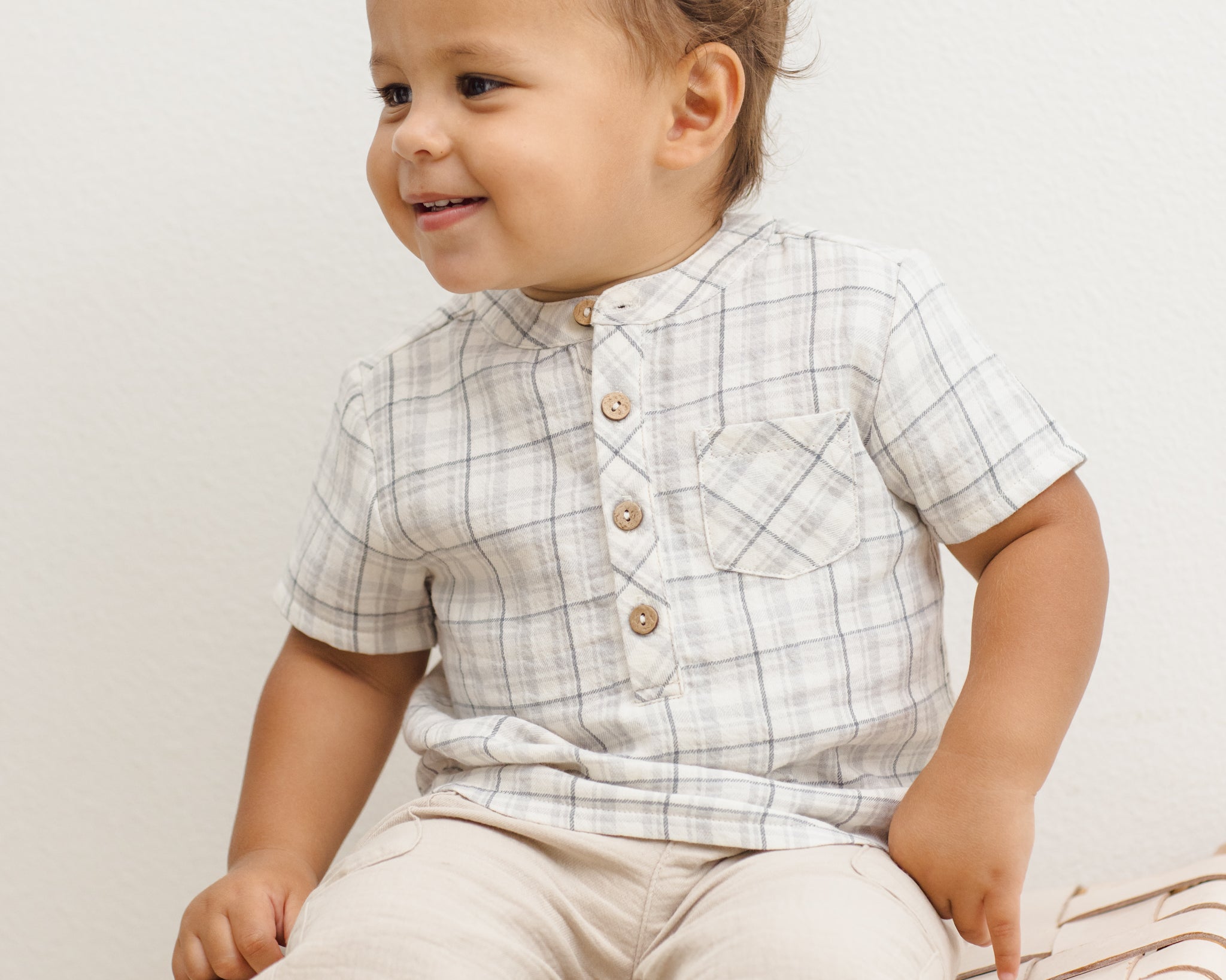 Toddler sitting on a rattan bench wearing the Rylee & Cru Jaxson Shirt in Blue Plaid