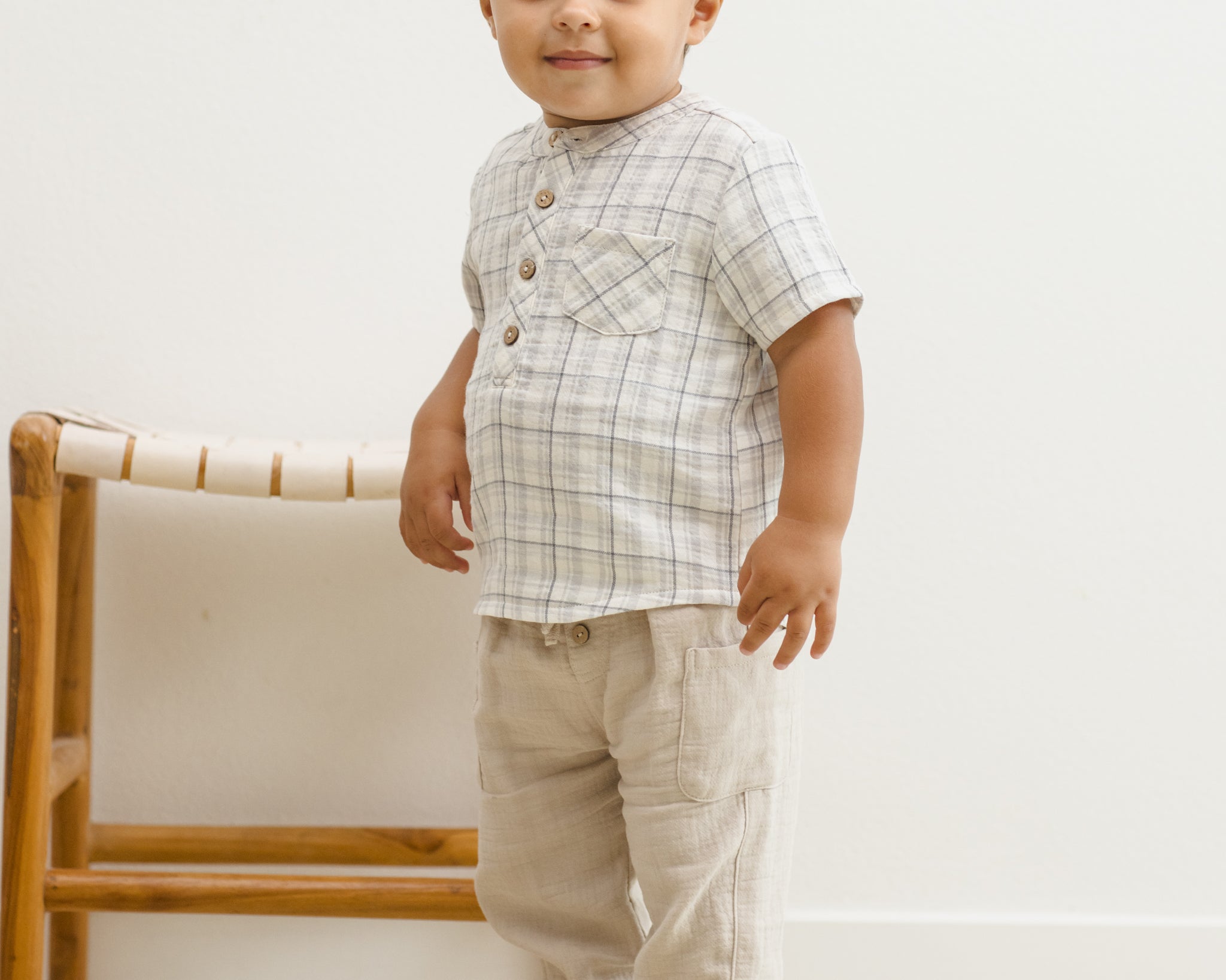 Toddler standing beside a rattan chair in the Rylee & Cru Jaxson Shirt in Blue Plaid and tan pants